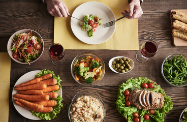 Top view of old male hands holding knife and fork and starting his salad. His dinner includes baked meat, spiced rise, carrots, green asparagus and vegetables