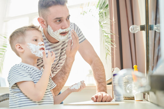 Handsome young man and his little son with shaving foam on their faces having fun together at spacious bathroom with panoramic window
