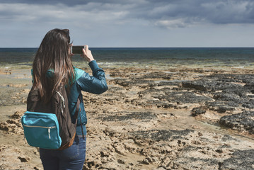 Back view of cheerful young woman photographing beautiful seascape while standing on rocky shore. She is using her smartphone. Copy space 