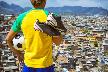 Brazilian football player standing in Brazil colors kit holding soccer ball in front of favela slum...