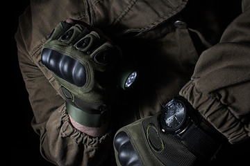 Photo of a male person in brown tactical outfit jacket and gloves using green tactical led flashlight and military watch. - obrazy, fototapety, plakaty