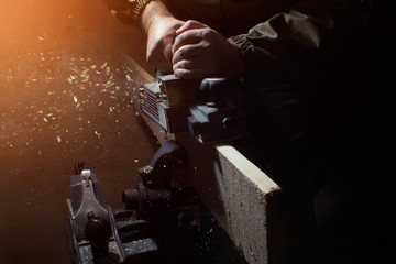 Photo of a worker processing a wood with electric plane with an old steel plane standing on vise with a dark background.