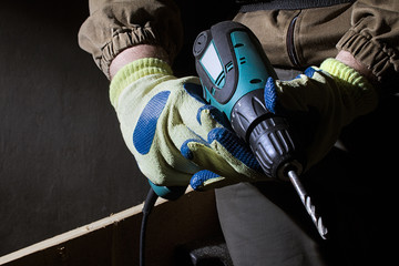 Closeup photo of a worker in outfit with blue gloves holding electric drill with wooden boards on...