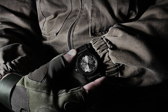 Profile view photo of a male person in brown tactical outfit jacket and gloves holding military watch on black background.