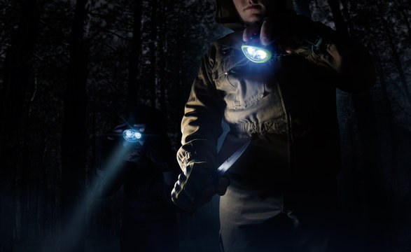 Photo of a two male person in brown tactical outfit jacket, gloves using knife and lighting up the way with head flashlights on night woods background.