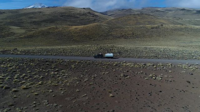 Aerial drone scene of van and trailer, motorhome in steppe, patagonia argentina rideing on a gravel lonely road. Tromen volcano. National Park. Camera moving left and going upwards tracking car.
