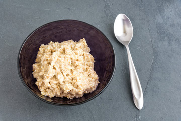 Fresh cooked oatmeal in a purple glass bowl with a metal spoon on a slate background

