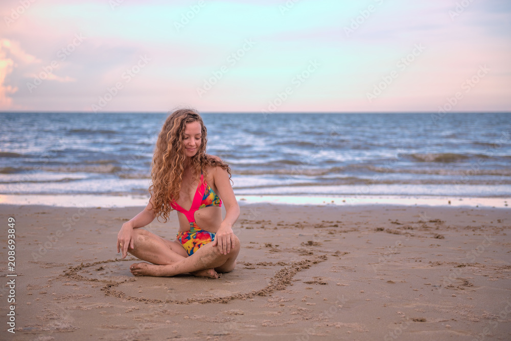 Wall mural young healthy woman with blonde curly hair sitting in a hand writing of heart shaped on the sand, su