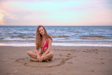 Fototapeta na wymiar Young healthy woman with blonde curly hair sitting in a hand writing of heart shaped on the sand, summer beach by the sea with waves, blue sky, relaxing and happy time concept.