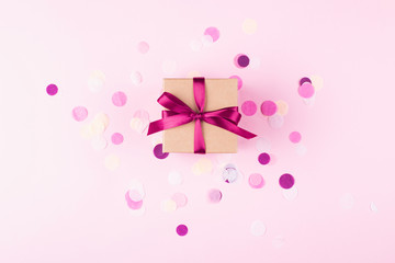 Kraft gift box tied with pink bow on pink background decorated with confetti.. Top view, holiday present concept.