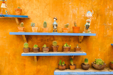 Many cactus and ceramic doll decorated on blue color cement shelf with orange wall.
