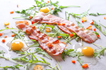 Tasty fried bacon, arugula and raw eggs on light background