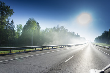 Asphalt road with metal safety barrier near the forest with thick fog over the sun.