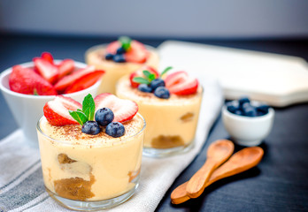 homemade, exquisite dessert tiramisu in glasses decorated with strawberry, blueberry, mint on black wooden table