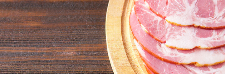 Sliced ham with fresh green lettuce leaves on a round cutting board. Meat products on a brown wooden table. banner