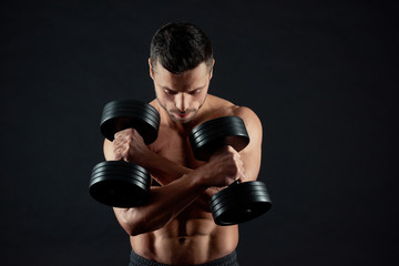 Portrait of athlete with strong muscular body keeping heavy metallic dumbbells with his hands crossed. Looking down, training hard. Having clear ABS relief, fit, athletic, sporty body.
