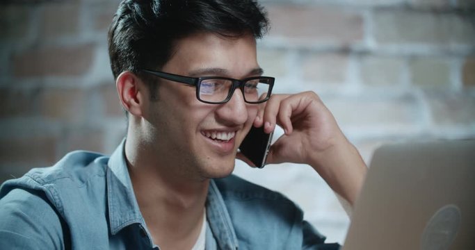 young handsome indian freelancer smiling while talking on phone and working on laptop dollyshot 4k