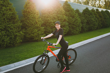 Young athletic brunette strong woman in black uniform, cap stop riding road on black bicycle with orange elements outdoors on spring or summer sunny day. Fitness, sport, healthy lifestyle concept.