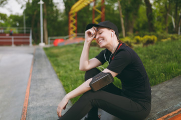 Young athletic relaxing girl with closed eyes in black uniform with headphones listening music, resting and sitting before or after running, training in city park outdoors. Fitness, healthy lifestyle.