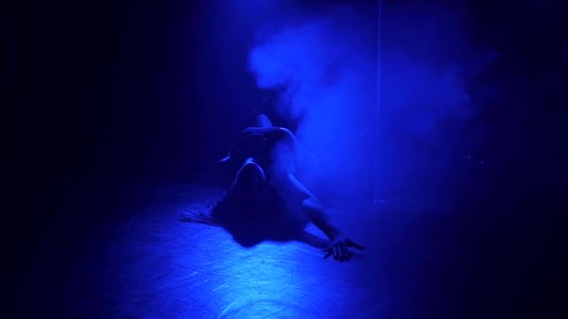 The silhouette of a young seductive girl dancing on the floor near a pole in the dark under the light of blue spotlights. Slow motion. Nightclub. Pole dance.
