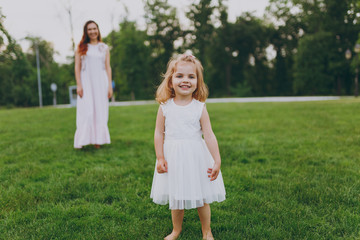 Naklejka na ściany i meble Smiling little cute child baby girl in light dress walking, play and have fun on green grass lawn in park. Mother, little kid daughter. Mother's Day, love family, parenthood, childhood concept.