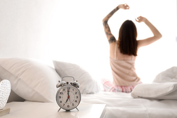 Table with alarm clock and young woman on bed in morning