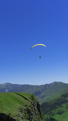 A paraglider with people flies over the mountain peaks in a bright blue sky.