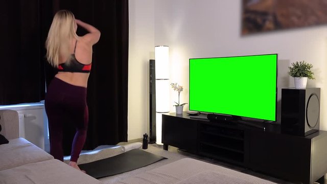 A young beautiful woman does a fitness dance in front of a TV with a green screen in an apartment