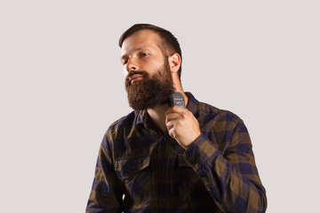 Making his beard perfect. Confident young bearded man shaving with electric razor and looking at camera in checkered shirt while standing against grey background