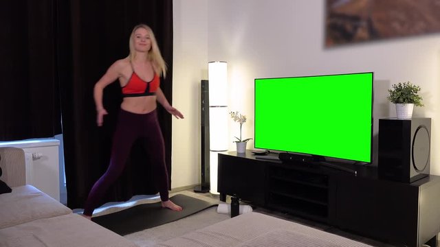 A young beautiful woman does a fitness dance in front of a TV with a green screen in an apartment and smiles at the camera