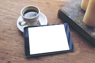 Top view mock up image of black tablet with white blank desktop screen and coffee cup , book , candles on vintage wooden table in cafe