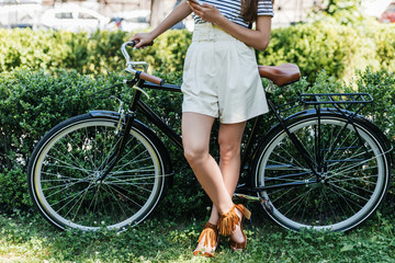 cropped shot of woman with smartphone and retro bicycle standing in park