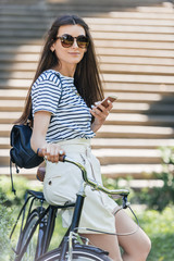 side view of attractive smiling woman with smartphone leaning on bicycle on street