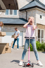 little kid covering eyes on kick scooter and her parents unpacking cardboard boxes for relocation into new house