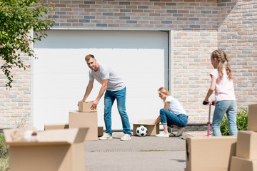 young couple unpacking cardboard boxes while their daughter riding on kick scooter in front of new cottage