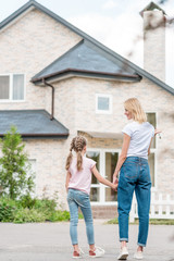 Back view of little daughter with mother holding hands in front of new house