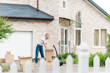 distant view of young woman unpacking cardboard boxes near guitar in front of new house