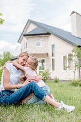 little daughter hugging smiling mother on green lawn in front of cottage