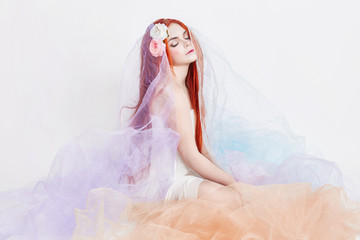 Redhead girl in light airy colored dress sits on the floor white background. Beautiful flowers in...