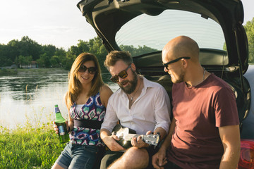 Group of happy people in a car at sunset in summer. Man sound ukulele and sing whit friends in the river at sunset. Romantic serenate.