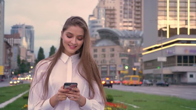 Young beautiful girl is typing message on her smartphone at sunset in city center in summer, communication concept