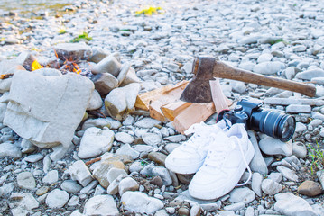 camp fire with fire woods and ax near it on sunset. white sneakers with dslr camera