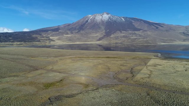 Tromen volcano in national park, Patagonia. Aerial scene moving right and down. Mountain with bed of lava and snow. Tromen lagoon with refletion in front of the volcano. Solitary gravel road