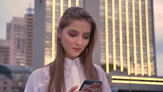 Young attractive concentrated girl is watching at her smartphone at sunset in summer, communication concept
