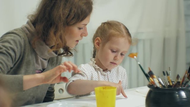 Children girls sit together at the table in the classroom and drawing with their fingers and paint. With them their young and beautiful teacher.