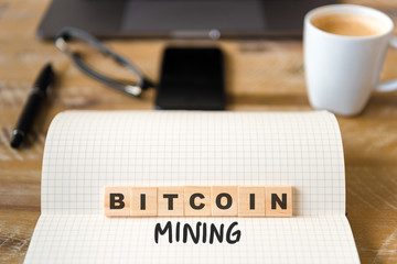 Closeup on notebook over vintage desk background, front focus on wooden blocks with letters making Bitcoin Mining text