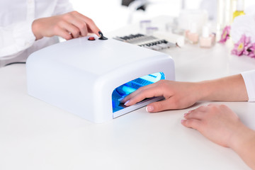 cropped image of woman holding hand in uv lamp while manicurist pushing button at table in beauty salon