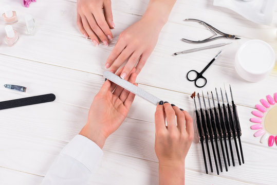 cropped image of beautician doing manicure by nail file to woman at table with cream, nail polishes, scissors, cuticle pusher, nail clippers, samples of nail varnishes