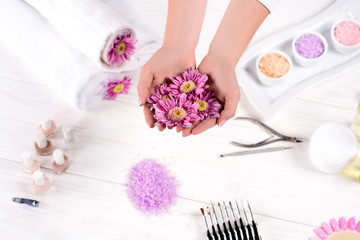 Obraz na płótnie Canvas cropped image of woman holding flowers over table with towels, nail polishes, colorful sea salt, cream container and tools for manicure in beauty salon