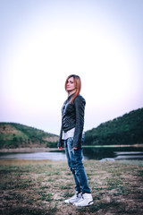 young woman in a leather black jacket and white shoes, jeans vintage jeans outdoor background, green hiils and river, sunset time